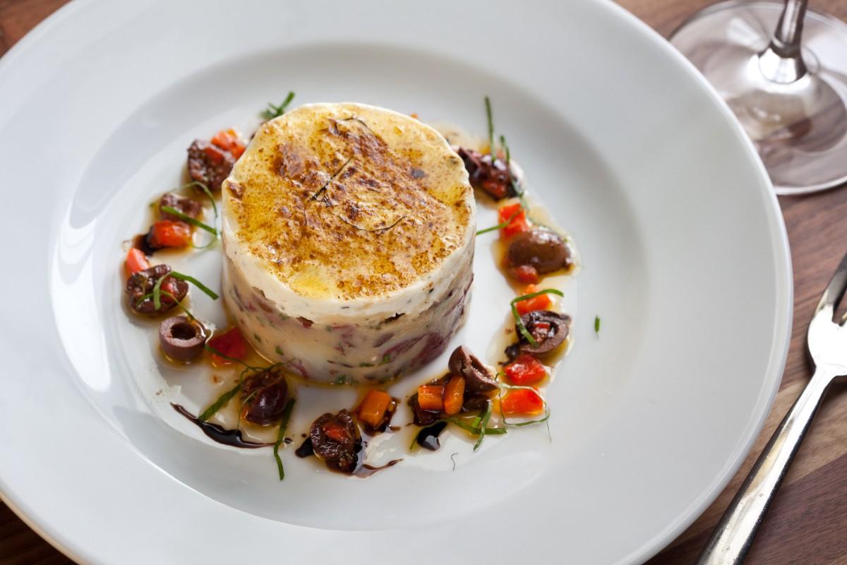A gourmet dish featuring a cylindrical molded salad or terrine, likely made of potatoes and finely diced vegetables or meat, topped with a golden-browned cheese or cream layer. It is garnished with a ring of chopped olives, red bell peppers, and fresh herbs, drizzled with olive oil and balsamic glaze, all presented on a white plate.